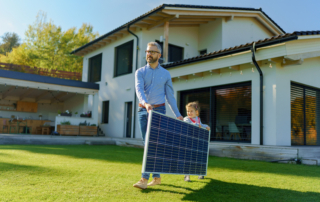 A father and young child carry a solar panel across the lawn outside their home.