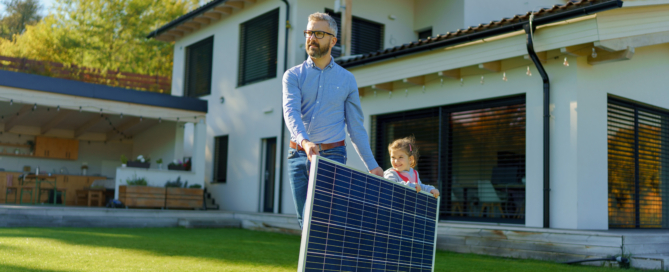 A father and young child carry a solar panel across the lawn outside their home.